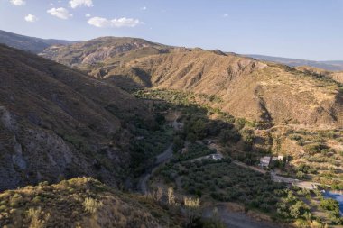 Yator 'un (Granada) yanındaki vadide, zeytin ağaçları, çalılar, vadiden geçen bir nehir var, gökyüzü açık.