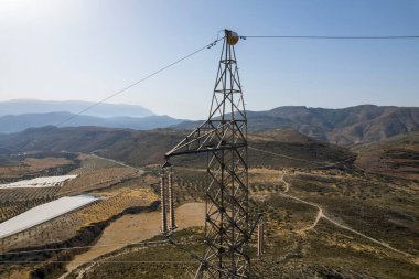 Granada dağının tepesinde yüksek voltaj kolonu var, sütunun tepesinde bir küre var, seralar var, tepelik bir alanda çalılar var, gökyüzü açık.