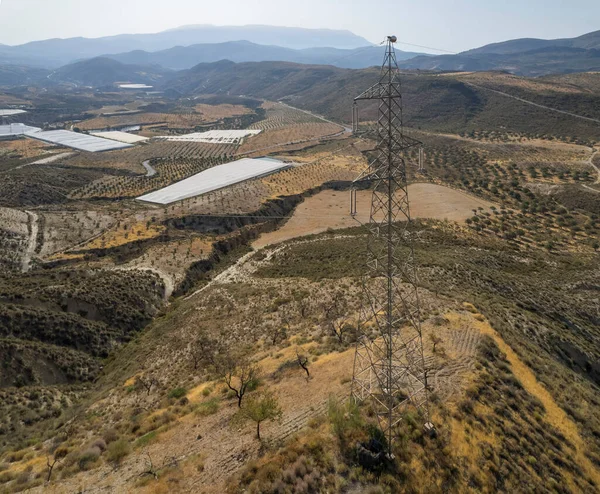 Granada dağında yüksek gerilim kolonu var, birkaç vadi var, seralar var, dağlarda ağaçlar var, gökyüzü açık.