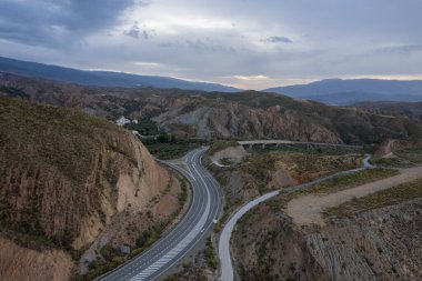 Granada 'nın güneyindeki dağların arasından geçen bir yol, beyaz çizgiler ve bir köprü var, dağların çalıları ve ağaçları var, gökyüzü bulutlu