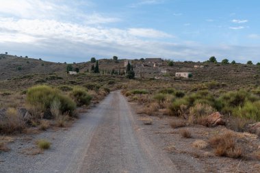 Terk edilmiş eski maden binalarına giden toprak yol, bir dağ alanı, çalılar ve ağaçlar var, gökyüzünde bulutlar var.