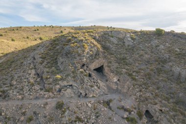 İspanya 'nın güneyindeki dağlık arazide toprak bir yol, bir mağara, dağlarda çalılar ve ağaçlar, gökyüzünde bulutlar var.