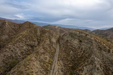 Dağdan geçen toprak yol, dağda çalılar var, birkaç vadi var, gökyüzü bulutlu.