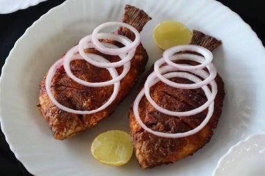 Çiğ soğan halkaları ve limon dilimleriyle süslenmiş beyaz bir tabakta servis edilen geleneksel Güney Hint balık kızartması. Munnar 'da yakalanan bu resimde bölgesel Kerala mutfağı sergileniyor, çıtır çıtır dokular ve yerel tatlar vurgulanıyor