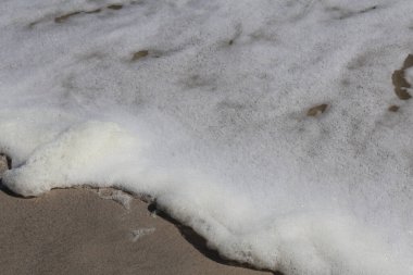 Beyaz Okyanus Köpüğü yavaşça Sandy Shoreline 'ın üzerinde süzülüyor. Köpüklü beyaz dalgalar kumlu bir sahile vuruyor ve ıslak kumla buluşurken köpüklerin ve kabarcıkların karmaşık dokularını yakalıyor. Plaj, doğa ve kıyı dinginliği temaları için ideal..