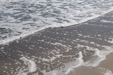 Beyaz Okyanus Köpüğü Sandy Sahili 'nde yuvarlanıyor. Islak kumun üzerinde süzülen köpüklü yumuşak dalgalar rahatlatıcı bir sahil manzarası yaratıyor. Plaj, okyanus ve doğa temaları için mükemmel. Deniz kenarının sakinliğini gözler önüne seriyor..
