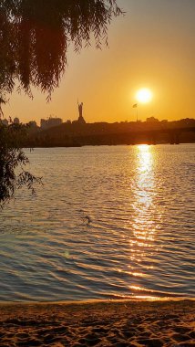 Nehre yansıyan altın renkleri ile nefes kesici bir günbatımı, bir heykel silueti ve uzakta bir köprüyü içeriyor. Huzurlu su ve ağaç dalları manzarayı çerçeveler.