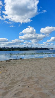 Sakin bir kumlu nehir kıyısı sahnesinde durgun suyun üzerindeki beyaz kabarık bulutların yansımaları, parlak mavi gökyüzünün altında şehrin ufuk çizgisi..