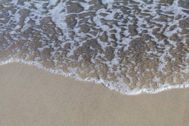 Köpüklü bir okyanus dalgası kumlu kıyı şeridiyle nazikçe buluşur. Güneş ışığında karmaşık desenler ve dokular yaratır. Kıyı ya da doğa temaları için mükemmeldir..