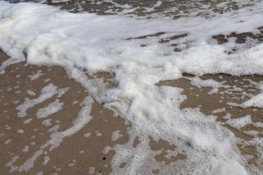 Kumlu sahil boyunca yayılan köpüklü deniz köpüğünün detaylı bir görüntüsü, sahil şeridinin doğal dokusunu ve hareketini yakalıyor..