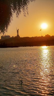 Anavatan Anıtı ve Ukrayna bayrağının dingin bir günbatımı görüntüsü, Kyiv 'deki dalgalı Dnipro Nehri' nin üzerine yansıyor, ağaç dalları tarafından çerçevelenmiş..