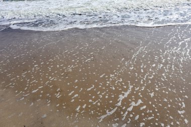 Köpüklü okyanus dalgaları yavaşça kumlu sahile ulaşıyor, sakin ve desenli bir deniz manzarası yaratıyor.