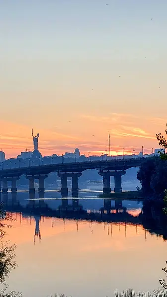 Ukrayna 'nın Kyiv kentindeki Dnipro Nehri üzerinde çarpıcı bir günbatımı, Anavatan Anıtı, bir köprü ve sakin suya yansıyan canlı gökyüzü yansımaları.