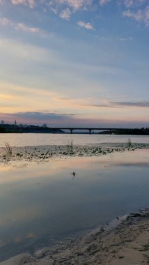 Kyiv 'deki Dnipro Nehri üzerinde sakin bir günbatımı manzarası. Huzurlu su yansımaları, bir köprü ve alacakaranlıkta alacakaranlığın yumuşak renkleri..