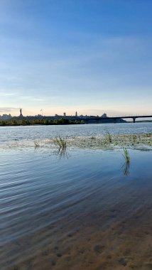 Dnipro Nehri 'nin sakin manzarası Kyiv siluetinin yansımaları, Anavatan Anıtı ve açık mavi gökyüzünün altındaki bir köprü.
