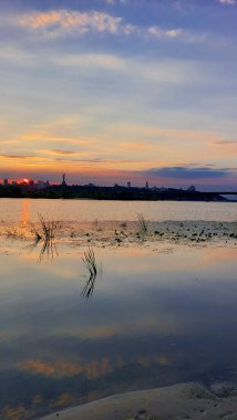 Huzurlu Günbatımı Dinyeper Nehri üzerinde Kyiv Skyline ve Anavatan Anıtı Yansıması ile. Alacakaranlıkta sakin bir nehir manzarası. Canlı gökyüzü renkleri, sazlıklar ve sakin su şehri yansıtıyor.