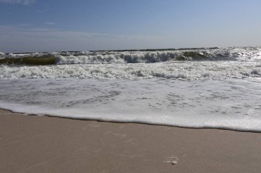 Güneşli bir gökyüzünün altında kumsalda okyanus dalgası köpüğü. Sahile vuran köpüklü dalgaların yakın çekimi, kumlu plaja karışıyor, parlak güneş ışığı ve hafif dalga hareketi huzurlu bir sahne yaratıyor.