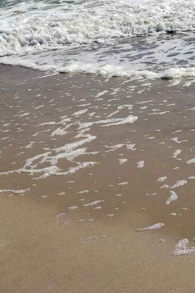 Deniz köpüğü desenleri kumsalda yakın planda. Geri çekilen dalgaların bıraktığı hassas köpük izlerinin detaylı bir görüntüsü kumlu kıyı şeridinin dokusunu ve güzelliğini vurguluyor.