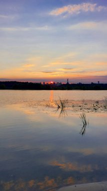 Sunset Over the Dnipro River with Kyiv Skyline and Reflection. A tranquil sunset scene with the glowing sun setting behind the skyline, reflecting on calm waters surrounded by reeds. clipart