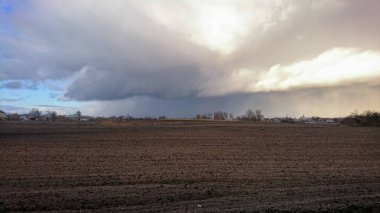 Fırtınalı bir gökyüzünün altında çarpıcı bir kırsal alan bulutlar ve yumuşak ışıkla çevrelenmiş uzak bir köyde soluyor, sakin ama dengesiz bir atmosfer yaratıyor..