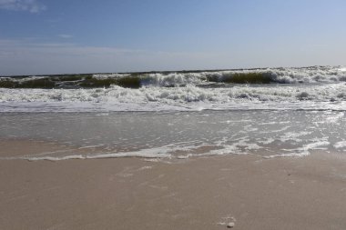 Açık mavi gökyüzünün altında, kumlu kıyılarda dalgaların dalgalandığı güneşli sahil, sakin ve sakin bir sahil sahnesini yakalıyor. Rahatlama ve doğa temaları için mükemmel..