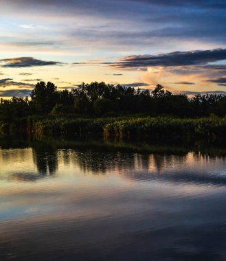 Nehrin üzerinde yaz günbatımı. Bulutlar ve ağaçlar nehre yansıyor. Sanatsal duvar kağıdı. Çok canlı bir atmosfer..