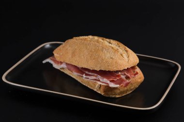 Koyu renkli tepside İber jambonlu sandviç, hepsi siyah arka planda açık kenarlı.