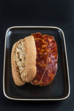 Sade bir kompozisyon ve siyah arkaplan içinde koyu renkli bir tepsiye yerleştirilmiş bir sucuklu sandviçin görüntüsü