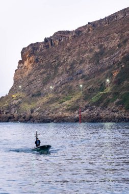 Yalnız bir balıkçı, gün batımında hafif ışıklarla aydınlanan dramatik uçurumlara karşı, sakin sularda küçük bir tekneyi yüzdürür. Pasajes San Juan, İspanya, Bask ülkesi.