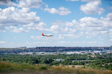 Bir uçak, bulutlarla dolu açık mavi bir gökyüzü altında şehir yapıları ve yeşil tepelere karşı sergilenen bir havaalanına doğru iniyor. Madrid, Barajas