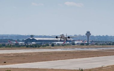 Kalabalık bir havaalanının pistinden büyük bir yolcu uçağı kalkıyor, açık bir günde arka planda bir hava trafik kontrol kulesi ve park edilmiş uçaklar görülüyor..