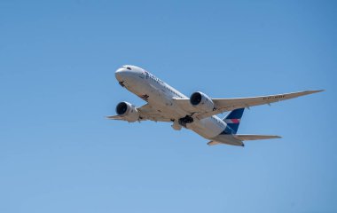 Büyük bir ticari uçak açık mavi gökyüzüne doğru kalkıyor, pisti terk ederken iniş takımlarını ve motorlarını gösteriyor. Latam Havayolları