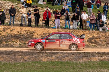 Mitsubishi Lancer WRC rali arabası toprak yolda hızla ilerliyor ve rotasında giderken toz topluyor. Seyirciler kenar çizgisinde toplanarak, yarışın açık havada gerçekleşmesini heyecanlı bir şekilde izliyorlar..