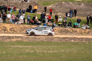 Ford Fiesta Rally3, dağların yamacında toplanan izleyicilerle dolu toprak bir patika boyunca hızla ilerlerken, kırsal alanda gündüz vakti meydana gelen heyecan verici motor sporu etkinliğinin tadını çıkarıyor..