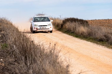 Kırsal alanda tozlu bir patika boyunca giden beyaz bir araba hareket ederken toz toprak ve toz topluyor. Yolu çevreleyen seyrek bitki örtüsü ve açık mavi bir gökyüzü var..