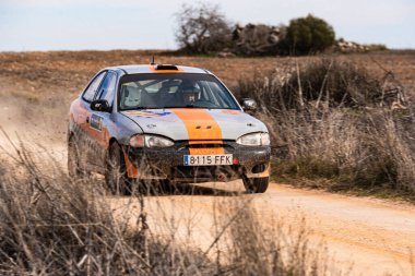 Hyunai Accent ralli arabası kırsal alanda tozlu bir yolda hızla ilerlerken, pistte giderken arkasında toprak bırakıyor. Araç parlak renkler gösterir ve yoğun bir odaklanma ile sürülür.