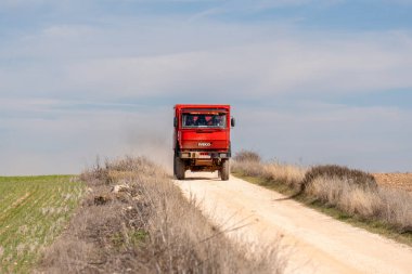IVECO engebeli kırmızı kamyonu, parlak mavi gökyüzünün altında canlı yeşil tarlalar ve yumuşak arazilerle çevrili tozlu bir yolda ilerliyor. Gün güneşli ve bulutsuz..
