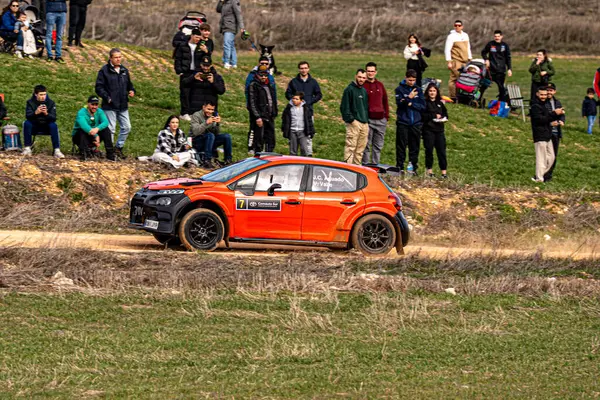 Citroen C3-R5 tipi kırmızı bir rali, güneşli bir günde yoğun motor sporu etkinliğini izleyen heyecanlı izleyicilerle çevrili bir toprak yolda hız yapar. Ortam tezahürat yapan hayranlarla dolu..