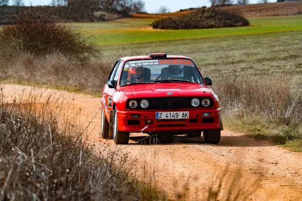 BMW serie 3 canlı kırmızı rali arabası yeşil tarlalar ve uzak tepelerle çevrili virajlı toprak bir yolda ilerliyor. Sürücü, kırsal bölgede hızla ilerlerken dikkatle odaklanıyor..