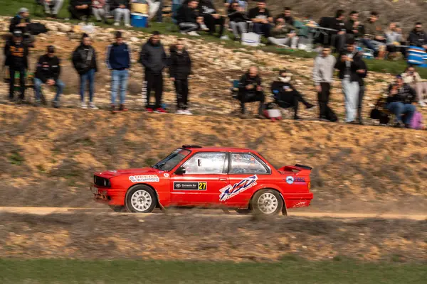 BMW serie 3 Enerjik bir rali arabası tozlu bir pistte hızla ilerlerken, büyük bir coşkulu seyirci kitlesi açık gökyüzünün altındaki bir yamaçtan izliyor..