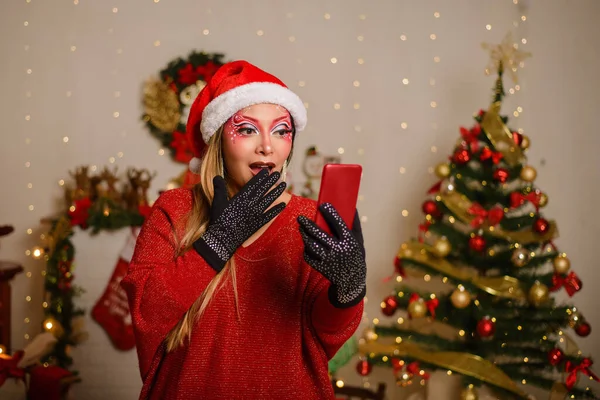 携帯電話を見ながら クリスマスのファンタジーメイクをした若い女性は驚きのジェスチャーを作ります クリスマスの背景 — ストック写真