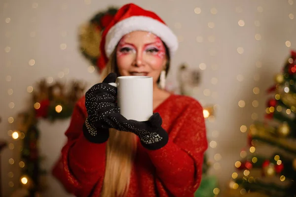 白いマグカップを持ってクリスマスファンタジーメイクを持つ若い女性 マグカップに焦点を当てます クリスマスの背景 — ストック写真