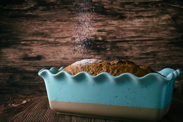 Pound cake in blue baking pan on wooden table. Sugar falling on the pound cake.