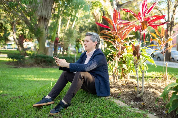 Gray-haired man looking at his cell phone sitting on the grass in a park.