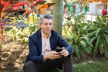 Gray-haired man looking at his cell phone sitting on the grass in a park.