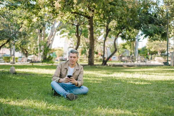 Homem Cabelos Grisalhos Olhando Para Seu Celular Sentado Grama Parque — Fotografia de Stock