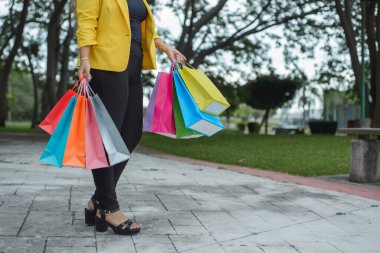 Sarı ceketli bir kadın farklı renkte alışveriş torbalarıyla bahçede yürüyor. Alışveriş konsepti.