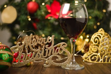 Ahşap masada bir kadeh kırmızı şarap ve Noel süslemeleri. Noel ağacı ve arka planda ışıklar.