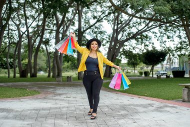 Sarı ceketli bir kadın farklı renkte alışveriş torbalarıyla bahçede yürüyor. Alışveriş konsepti.