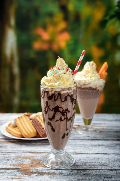 Chocolate milkshake with whipped cream decorated with sprinkles on a wooden table. Milkshake and sweet cookies with summer background.